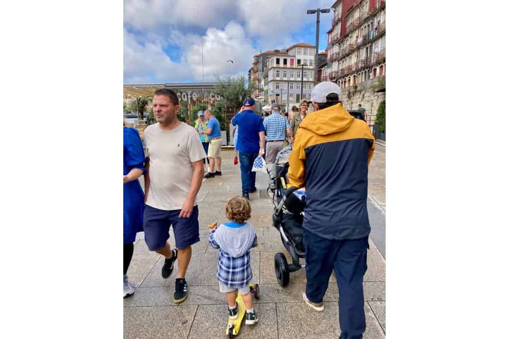 A man it pushing a pram along the waterfront in Porto with his young son next to him on a yellow scooter.  This is a day trip from Lisbon.