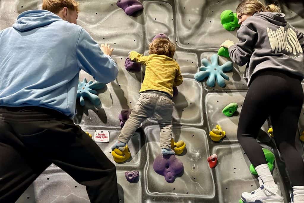 A boy in a yellow top and grey trousers is climbing a climbing wall with multicoloured hand holds.  On either side of the child is an adult, they are wearing jogging bottoms and hoodies and also climbing next to the boy.