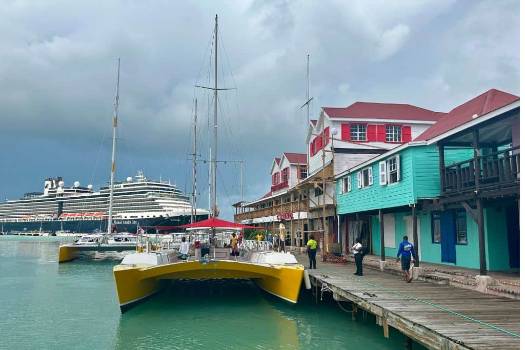 The blue, red and white historical buildings of Redcliffe Quay with a Wadadli Cat tour boat moored alongside it.  In the background is a large cruise ship at the Antigua cruise port.