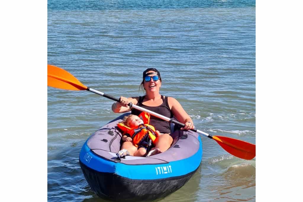 A woman in a blue inflatable kayak with her son between her legs in an orange life vest asleep.  They are paddling on the River Hamble which is a fun childrens attractions in Hampshire.