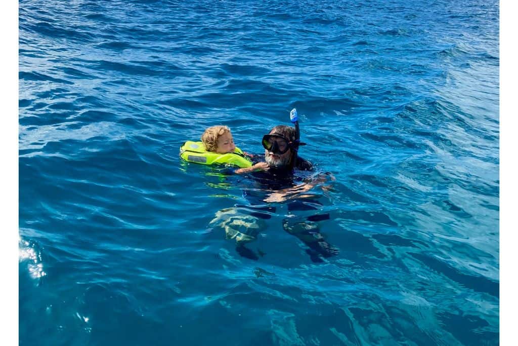 Boy in a yellow life jacket being held in the water by his dad who has a snorkel mask on at Cades Reef which has some of the best snorkelling in Antigua.