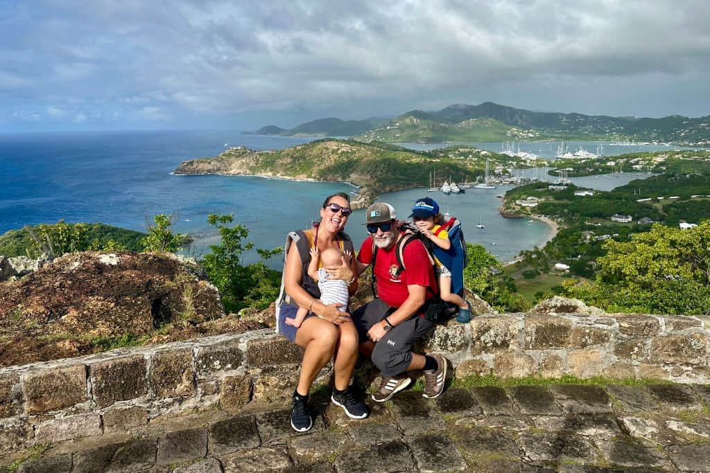 Family of four sat in front of the main view from Shirley Heights lookout.  In the background are the boats moored up in English Harbour.