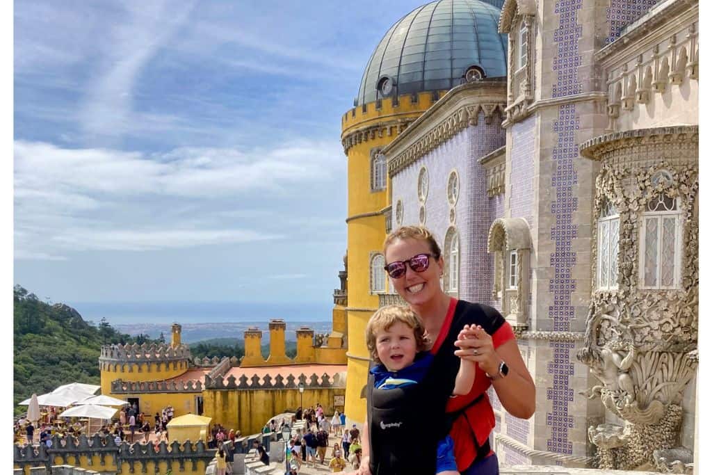 Mum wearing an orange t-shirt with her toddler son in a baby carrier with him facing outwards in March.  In the background is the Pena Palace which is close to Lisbon.