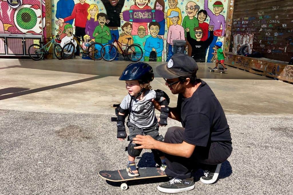 A two year old boy has a helmet on with knee and elbow protection and is stood on a skateboard.  Next to him is a man dressed in black with a baseball cap on teaching him to skateboard.  Learning to skate board with kids is such a fun things to do in Lisbon.