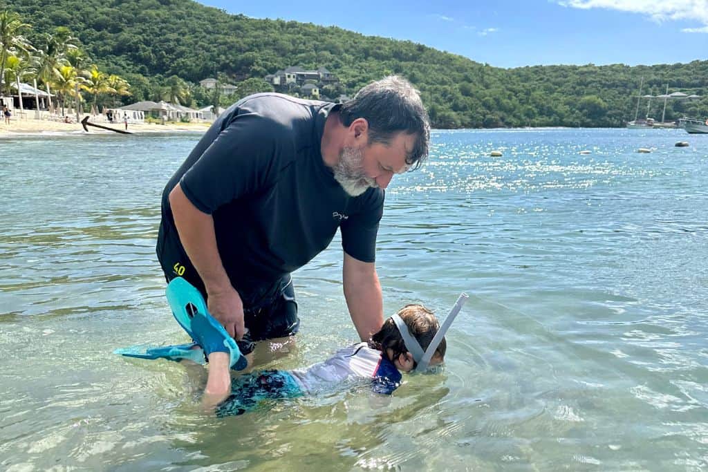 Father wearing a black UV t-shirt and swim shorts helping his son in the water to learn to snorkel in Dickenson Bay which is near to Antigua cruise port.