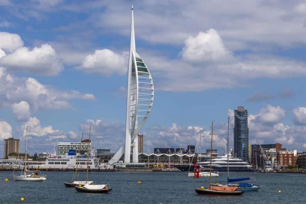 The view from the water of the front of the Spinnaker Tower in Portsmouth which is a sail shaped building. In front of it are some boats in the water.