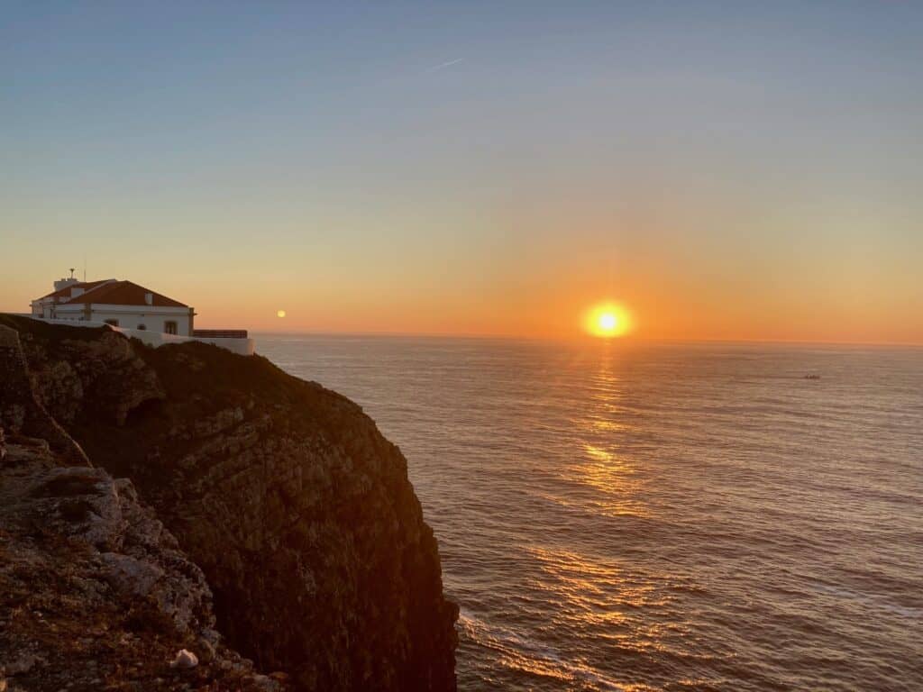 There is the edge of the cliff with a small white building on top of it on the left and then in the middle of the image is the sun setting into to the sea in Sagres in Portgual.  Something that should be on your bucket list.