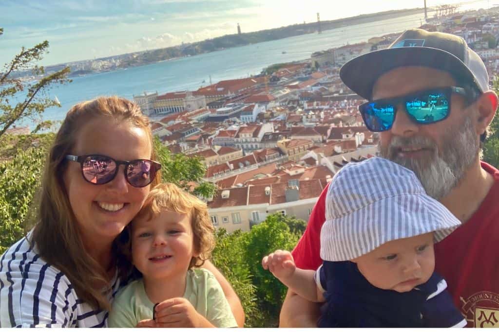 mum in stripey t-shirt, dad in red t-shirt with their young kids with the redroofed view of Lisbon behind them.  The view is from Castelo de São Jorge which is one of the best things to do with kids in Lisbon.