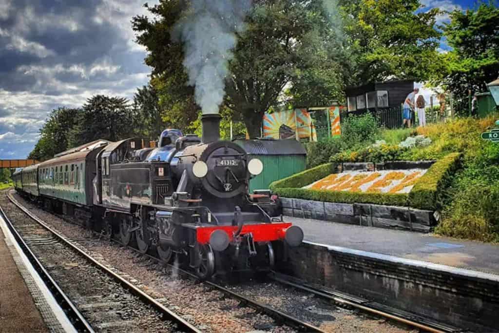 An old fashioned steam train with a black front section. The train appears to be moving as there is steam coming out of the train.  The train is on train tracks at The Watercress Line which is a fun childrens attractions in hampshire