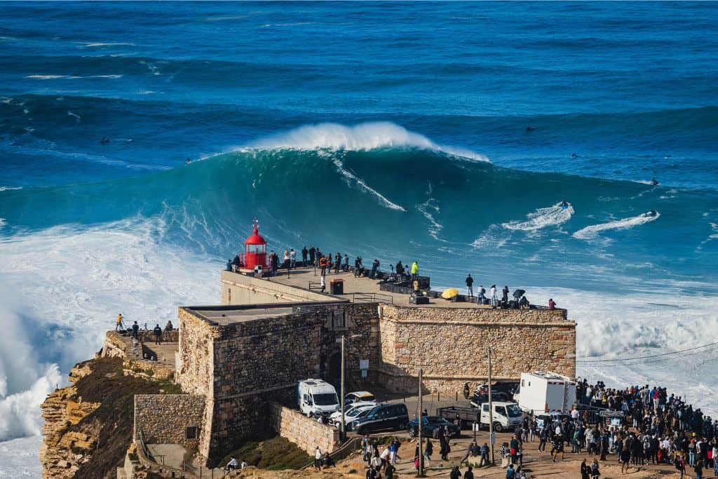 This is the fort with the red lightouse on top of it at Nazare and in the background there is a massive wave. 