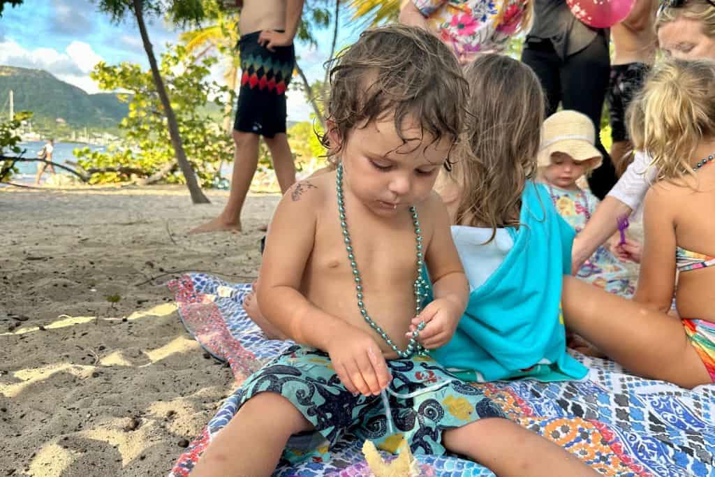 A little boy is sat on a blanket on the beach with some turquoise necklaces on with his friends in the background