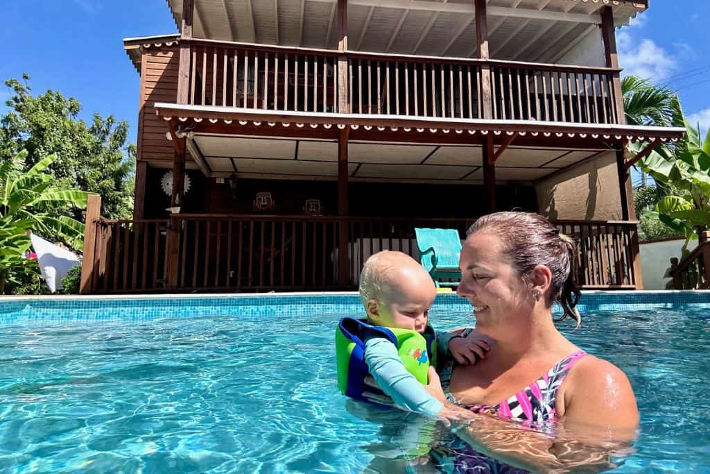 Lady in a swimming pool is in a colourful swimsuit holding her baby boy who is in a swim aid vest and is looking at his mum.  In the background is the house that they live in as a family. They are in Antigua as she is a digital nomad.