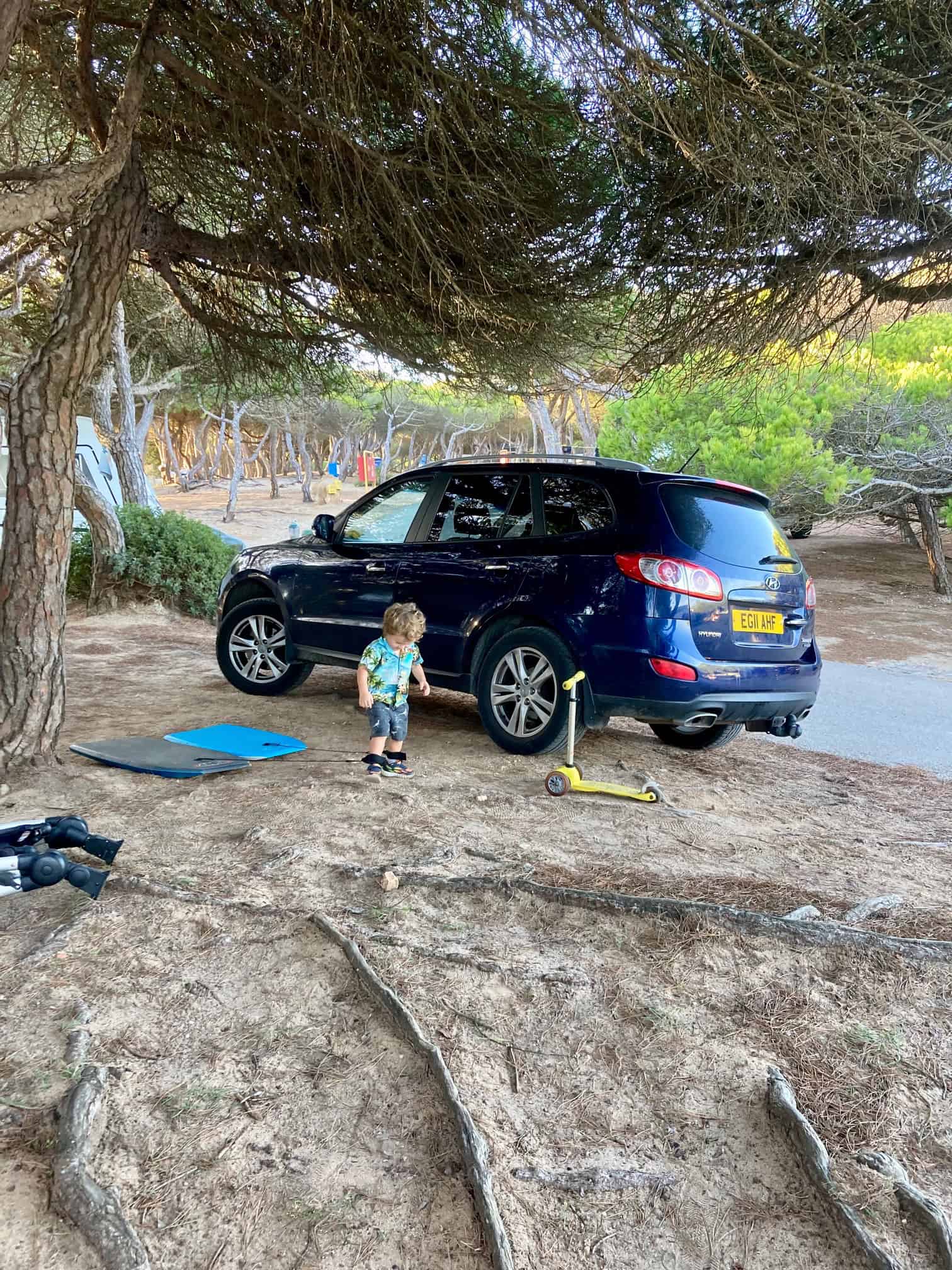 A blue car under some trees at a campsite in Lisbon. In front of the car is a little boy playing on his scooter.  They are about ot go to Nazare for a day trip from Lisbon.