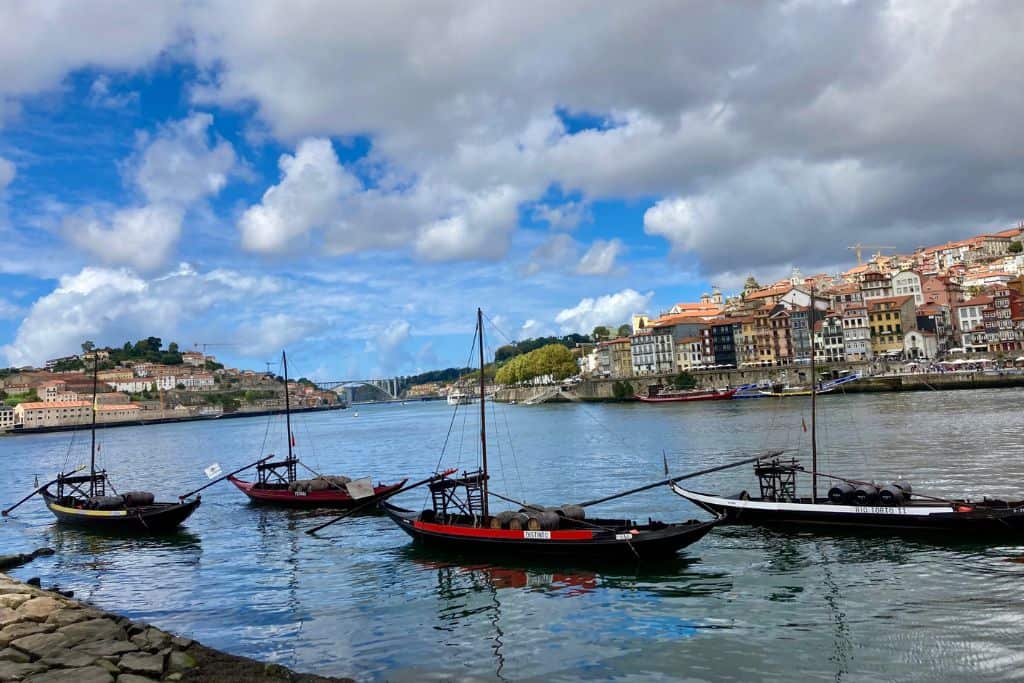 The Douro River in Porto, and on it are 3 historic small boats tht used to carry port barrels throughout the city.  On the far side of the river you can see some building on the waterfront.  In the blue sky there are lots of white clouds.