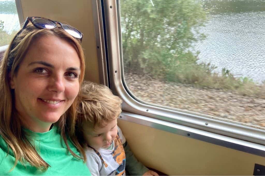 A woman is looking at the camera and smiling as she is sat on a train that's going along the Douro Valley from Porto. On her lap she has her toddler son who is looking out of the window at the scenery.