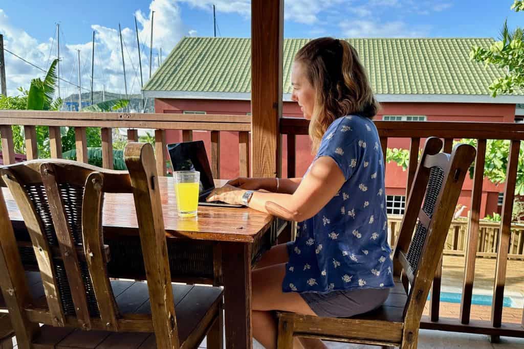 A lady in a blue shirt is sat at a dark wood table on her laptop working. She is in Antigua with her family and is a digital nomad.