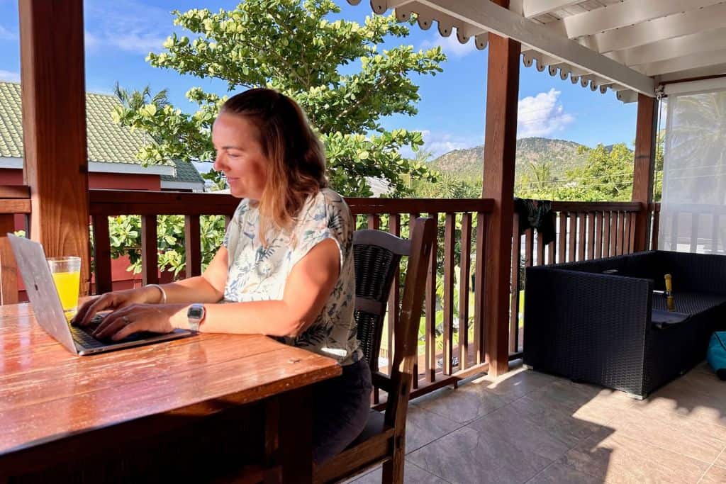 A lady in a light coloured flowery shirt is sat working on her laptop at a dark wood table in Antigua. She is a digital nomad and lives there with her family