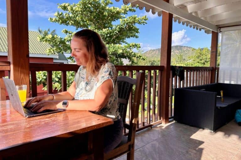 A lady working at a dark brown table on a balcony next to a pool. She is typing into her laptop and looking at the screen as she is a digital nomad in Antigua with her family.