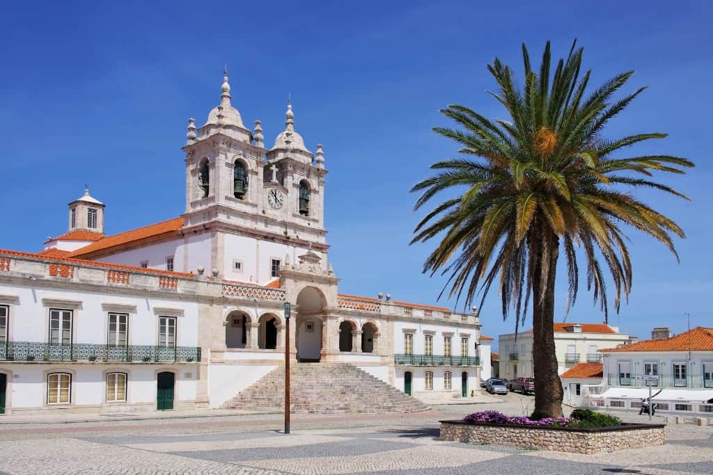 This is the outside of the Sanctuary of Our Lady of Nazare which is where they built a shrine to celebrate the legend of Nazare.  The building is white with two bell towers.  The roof is red and in front of the building is. palm free.