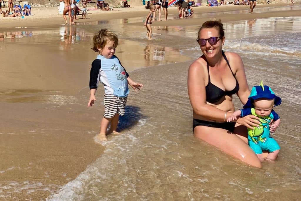 A woman is sat in the sand of a beach with small waves about an inch high coming in over her. On her lap is her 7 month old baby son in a wetsuit, and behind her is her toddler son laughing in his swimwear and about to enter the sea. She is travelling in Portugal with her kids. 