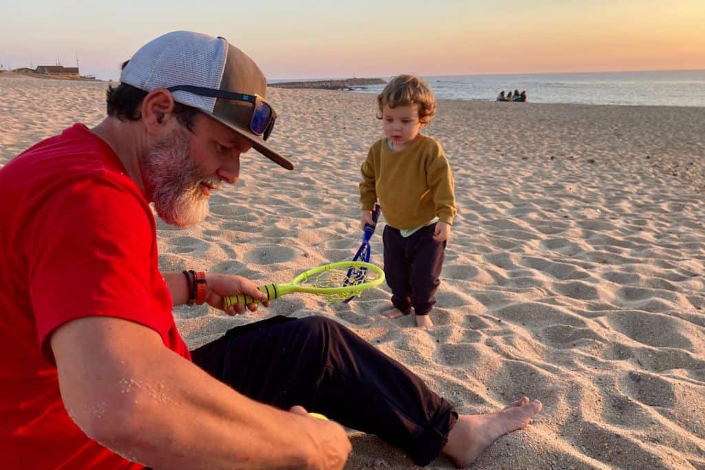 A man in a red t-shirt is sat on the beach and opposite his is his toddler son.  They both have tennis racquets in their hands and are plying a game of beach tennis whilst in the background the sun is setting. 