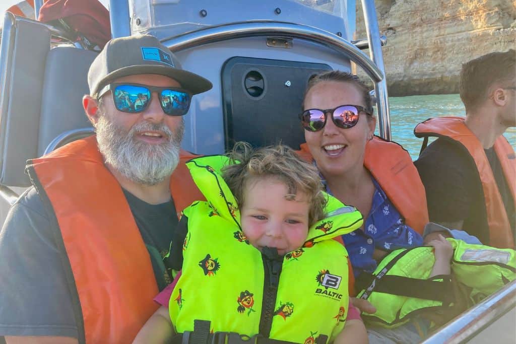 A family are sat on a bench sea on a boat about to visit the Benagil Cave in The Algarve.  The mum and dad are both wearing sunglasses and orange lift vests. Their 3 year old sone has on a bright yellow lifejacket with small pirates as a pattern on it.  The mum is holding a baby on her lap that also has a plain yellow lifejacket on. They are on a family trip to Portugal.