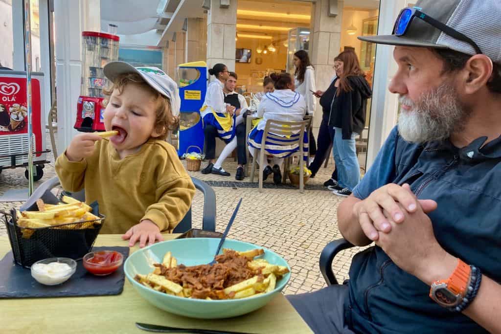 A father and son are sat at an outside cafe in Ericiera.  The toddler is eating a chip and the father is looking at him.  He is in Portugal with his kids.