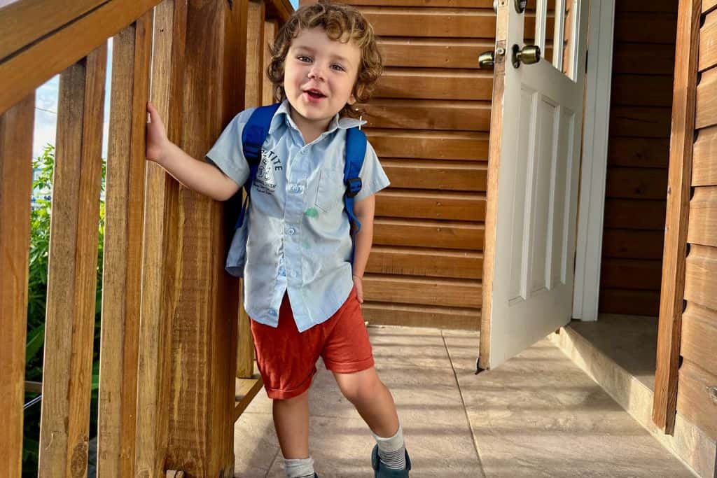 A little boy it standing on some stairs.  He has on a light blue school shirt and a small blue backpack as he laughs into the camera. He lives in Antigua with his family and is about to go to school as his mum is a digital nomad.