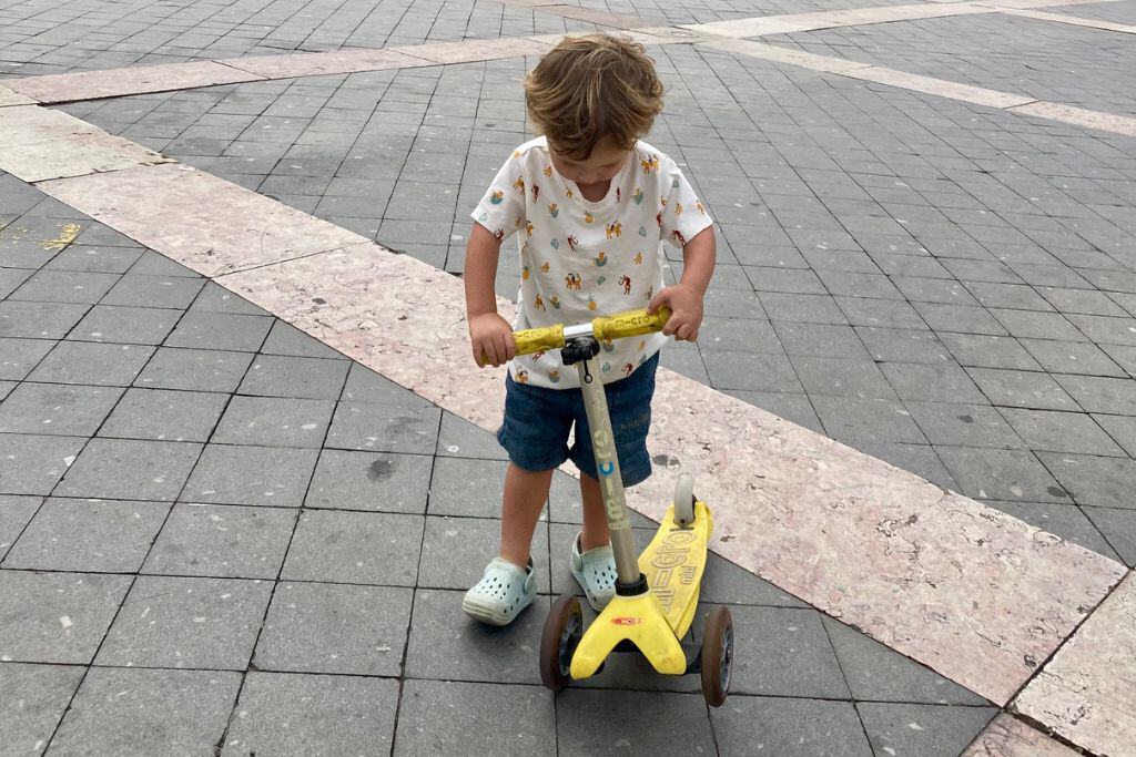 A 2 year old boy is stood on his yellow scooter looking at his shoes.  He's in Lisbon in Portugal.