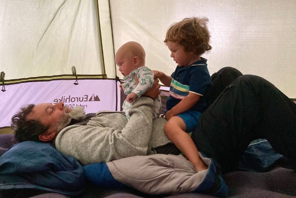 A dad is lying in his fleece on the floor of a tent and sat on him are his baby son in a colour onsie and his toddler son in blue shorts and a polo shirt with stripes on. He is in Portugal with his kids.