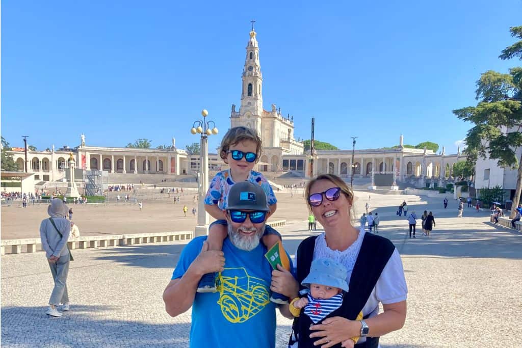 A man wearing sunglasses, a baseball cap and blue t-shirt is stood with his son sat on his shoulders.  His son hason sunglasses too.  Next to them is the mother in a white t-shirt, also wearing sunglasses and she has a baby carrier on her front.  They are stood in the main square at the Sanctuary of Fatima as they are on a family trip to Portugal.