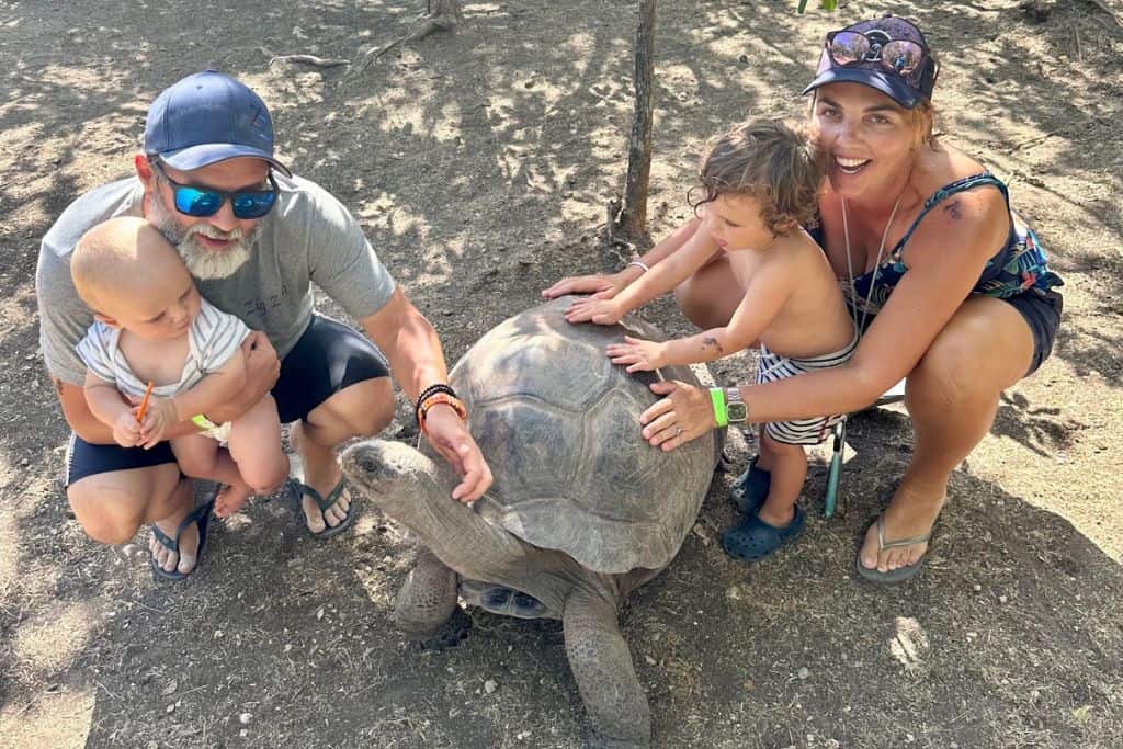 A man is holding his baby and is crouched down next a giant tortoise whilst he pets it.  Next to him is the mum with their other young son and they are also petting the giant tortoise. They are living in Antigua as a digital nomad family.
