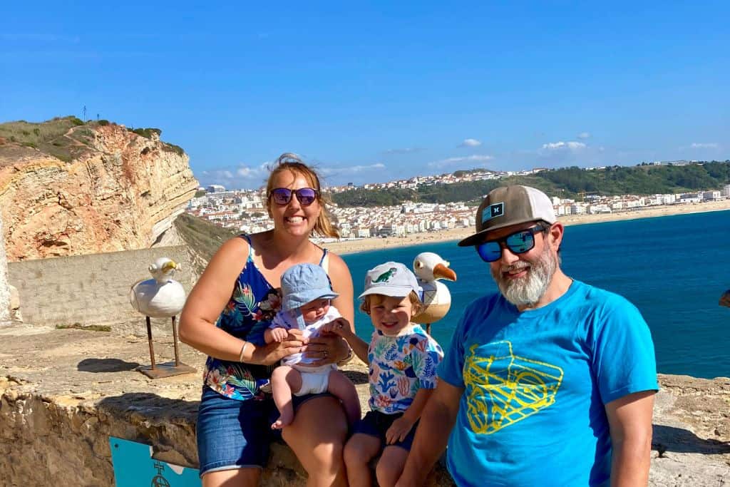 A mum, dad and gtheir two young children are sat on a wall outside the lighthouse in Nazare in Portugal. On the wall is a wooden sea gull.  In the background behind them is the beack with the blue sea and town centre. They are in Portugal with their kids.