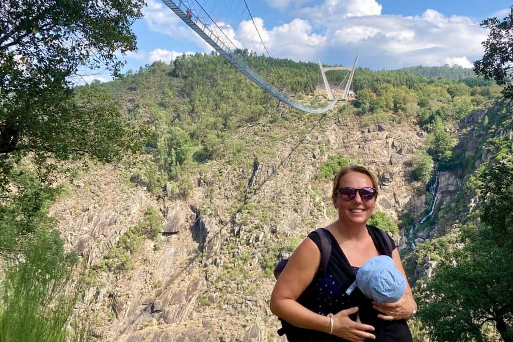 A mum wearing sunglasses is stood in front of Europes longest suspension bridge as part of the Paiva Walkways hike.  On her front is her baby in a baby carrier as she is on a family trip to Portugal.