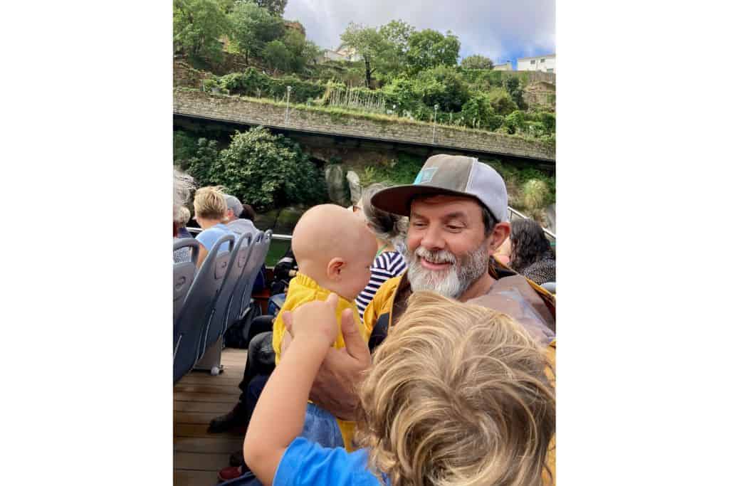 A dad is sat on a river boat wearing a baseball cap holding his son who is wearing a bright yellow jumper.  In front of them you can see the back of the 3 year olds head.  In the background is the sideview of Porto from the river.