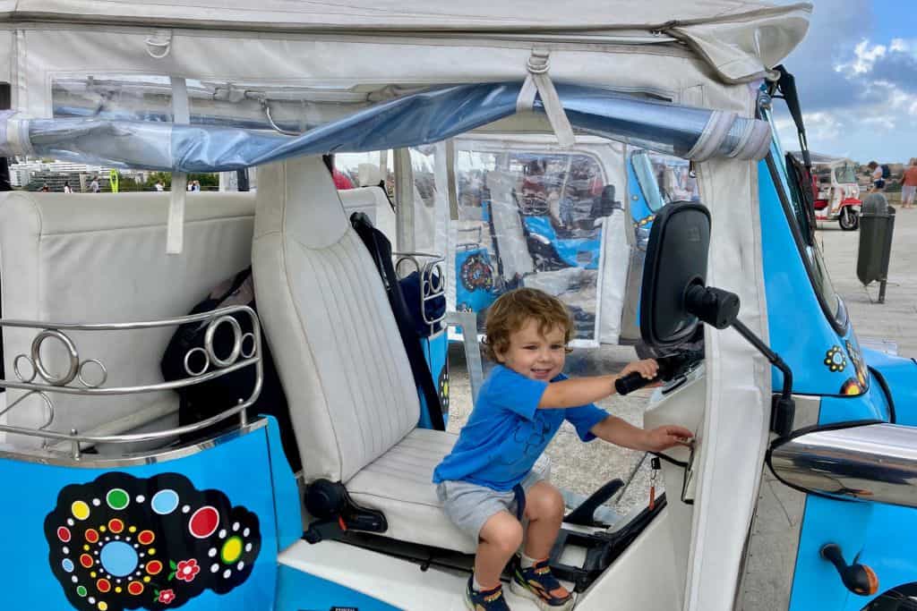 A toddler in a blue t-shirt and shorts is sat in the drivers seat of a blue tuk-tuk.  He has the keys and is looking at the camera with a cheeky smile pretending to start the engine.