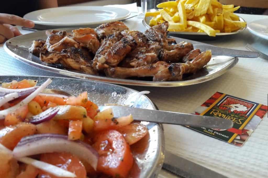 This is a close up on three dishes, on one of them is a tomato and onion salad, in the middle is a cut up whole chicken, and on the far side at the back of the image is a dish of chips.