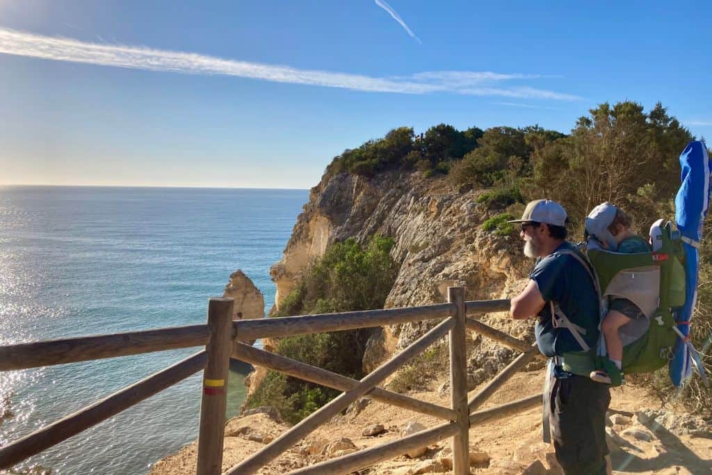 A man is stood resting his arms on a wooden railing at the edge of a cliff over looking the sea.  He is looking out at the sea and has his toddler son on his back in a special backpack carrier.  This is the Deven Hanging Valleys trail in The Algarve in Portugal and he is hiking it with his kids. 