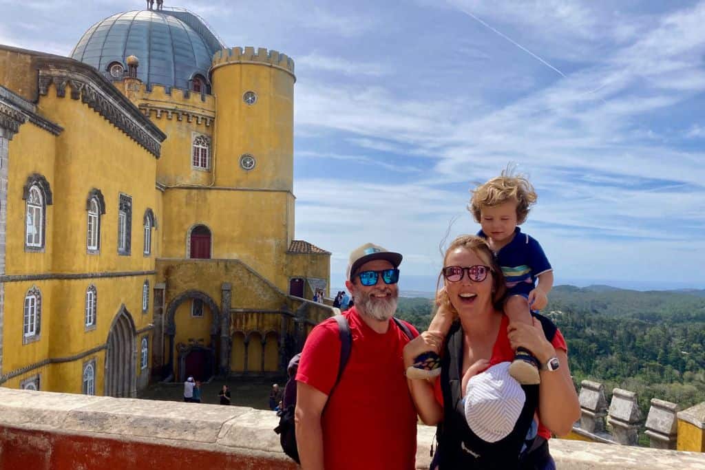 There is a family stood in front of the yellow main building of the Pena Palace in Sintra. The dad has on a red t-shirt, baseball cap and sunglasses.  The mum has on an orange t-shirt, and sunglasses.  On her front in a carrier is their baby. And on her shoulders is sat her 3 year old son who is wearing short and a polo shirt. They are on a family trip to Portugal