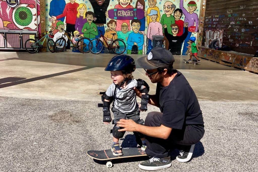 There is a boy in grey t-shirt and shorts, he has on knee, elbow and wrist pads and bike helmet and is stood on a skateboard that is facing the left of the picture.  Crouched next to him is a skater dressed in black with a baseball cap on who is teaching him how to skateboard. In the background is a large wall that has a big image of graffiti on it as they are in a skatepark.
