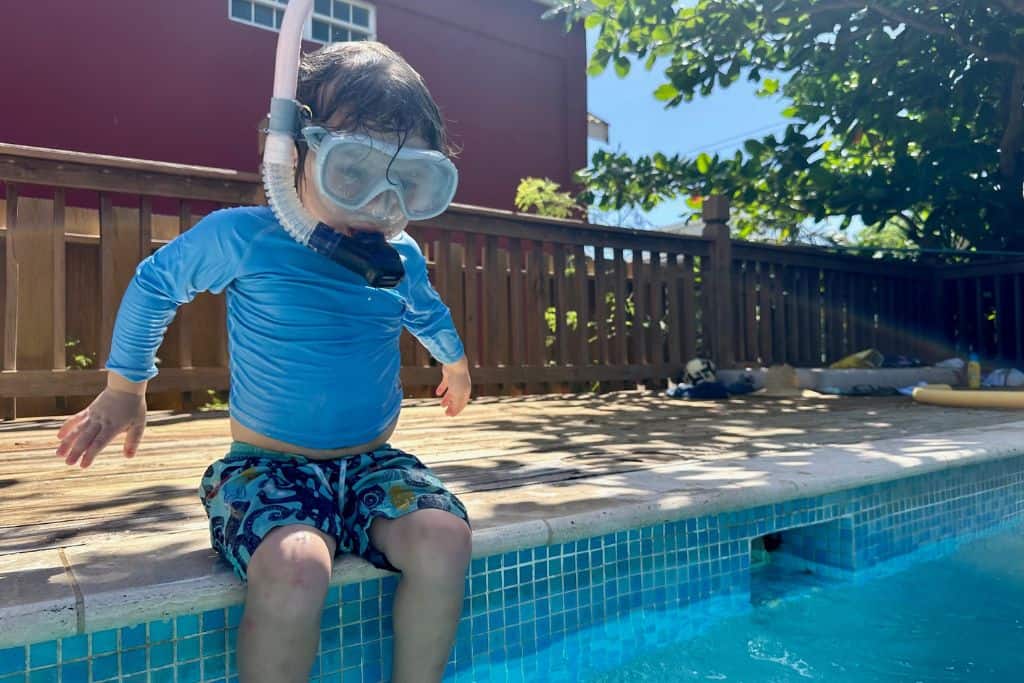 Young boy sat on the side of a swimming pool with his snorkelling gear on. His parents go snorkelling with kids.