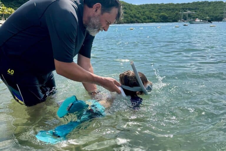 A man is stood in the shallow waters of a beach. He's holding the back of his son's UV t-shirt as he's practising to snorkel