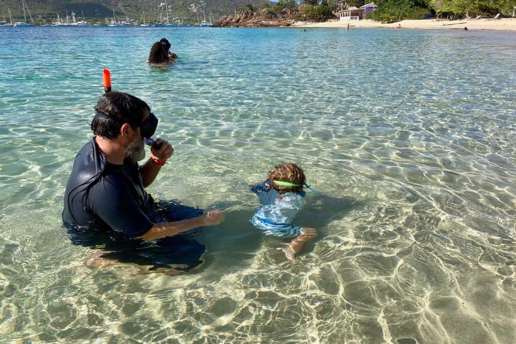 A man is crouched in the shallow water wearing s snorkel mask. His son is also crouched in the water and is walking along the beach with his face in the water. His dad enjoys snorkelling with kids.
