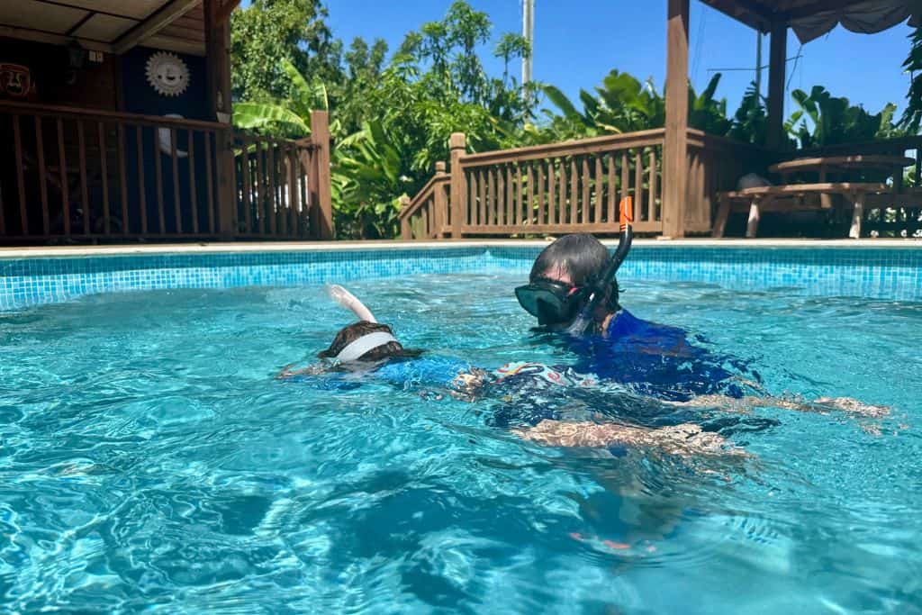A young boy is in his swimming wear in his swimming pool.  He's face down in the water with his snorkel mask on.  His dad is stood in the water next to him with his snorkel mask on, and is enjoying snorkelling with his kids.