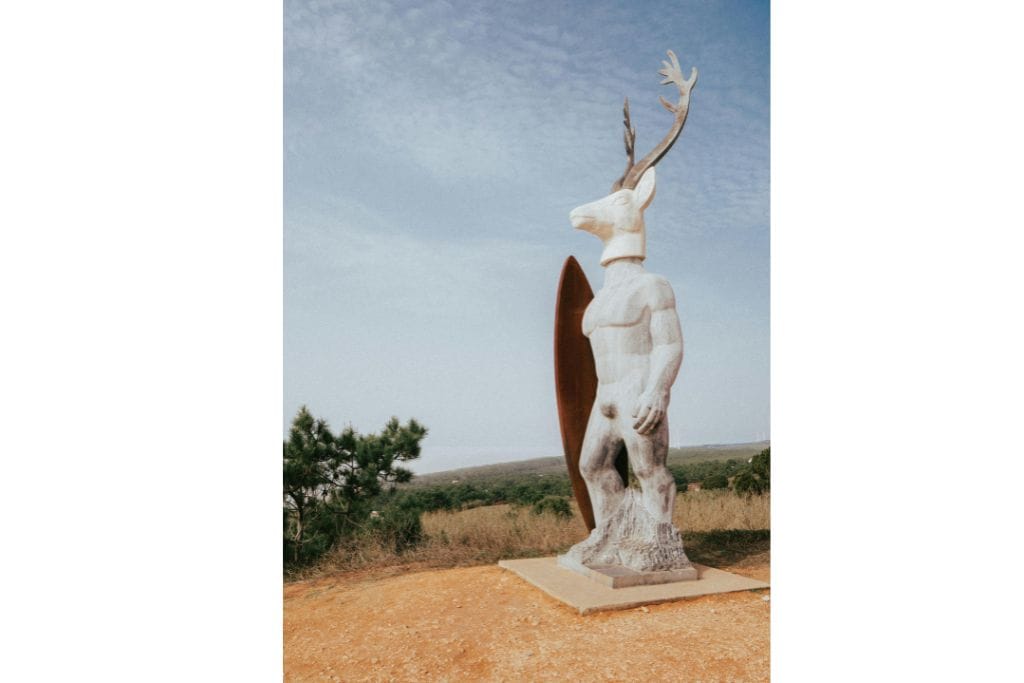 This is the statue in Nazare which is made of marble and steel.  The legs, body and arms are of a man and the head is a deer. In their arms they are holding a brown surfboard.