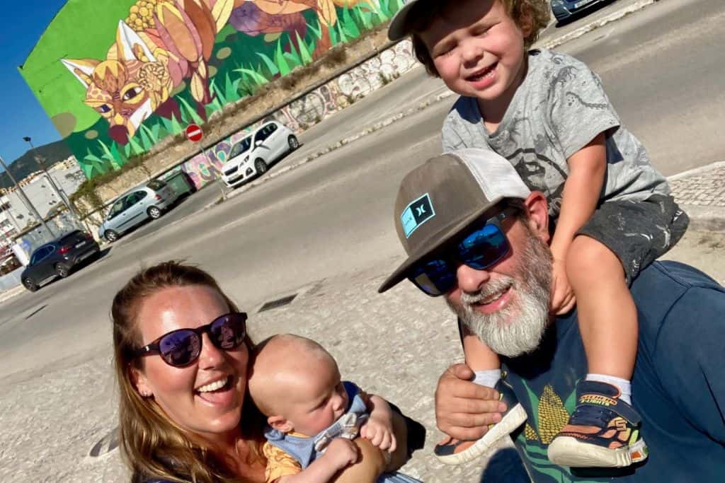 A woman is holding her baby, and next to her is her boyfriend who has their toddler son on his shoulders.  They are stood on the edge of a road and behind them is street art mural of a fox. They are travelling Portugal with their kids.