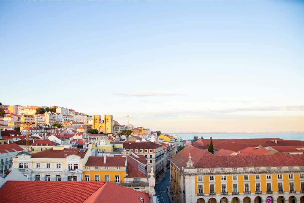 A city scape of Lisbon city centre.  In the image are a selection of buildings, some that are white and some that are yellow and they all have red roofs.  The sky is blue and there are no clouds.