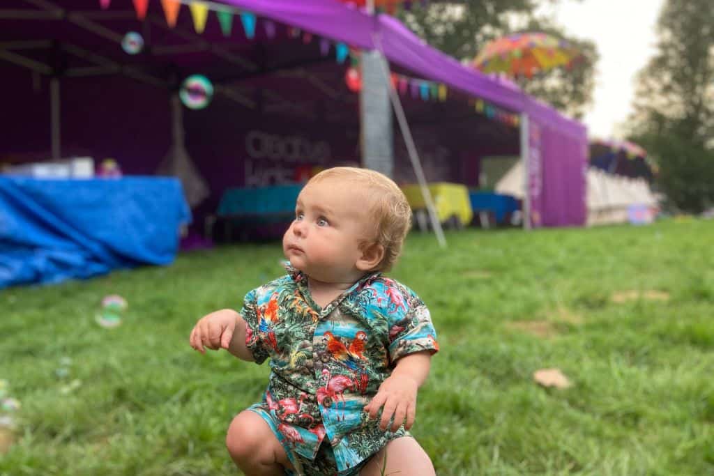 A baby in a Hawaiian outfit is looking to the side of the photo at some bubbles in the air. In the background is a marquee.  This is the Isle Of Wight Festival, one of the best festivals in Hampshire.