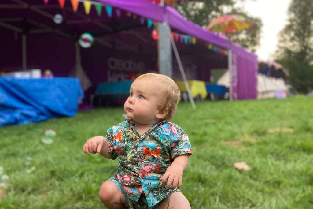 A baby boy is in a Hawaiian shirt and shorts sat on the grass looking off to the side as some bubbles re floating away