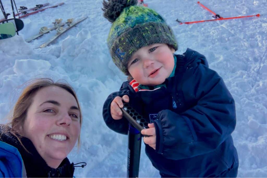 A mum is smiling at the camera sat in the snow and next to her is her two year old son leaning on a snow shovel in the snow.  They are both in their ski gear as she has been skiing with her kids.